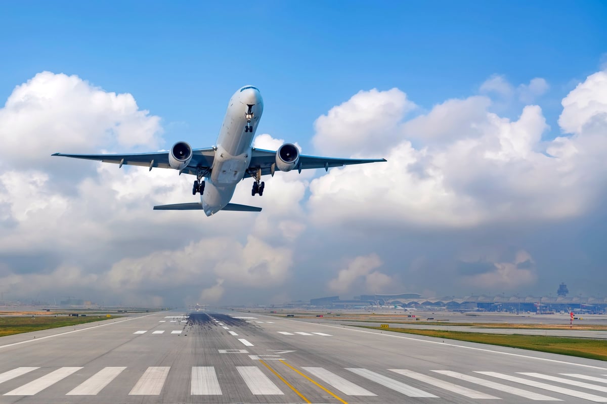 View airplane ascends into the sky after leaving the runway. The clear blue sky is dotted with white clouds, and an airport is visible in the background, showcasing a bustling travel scene.